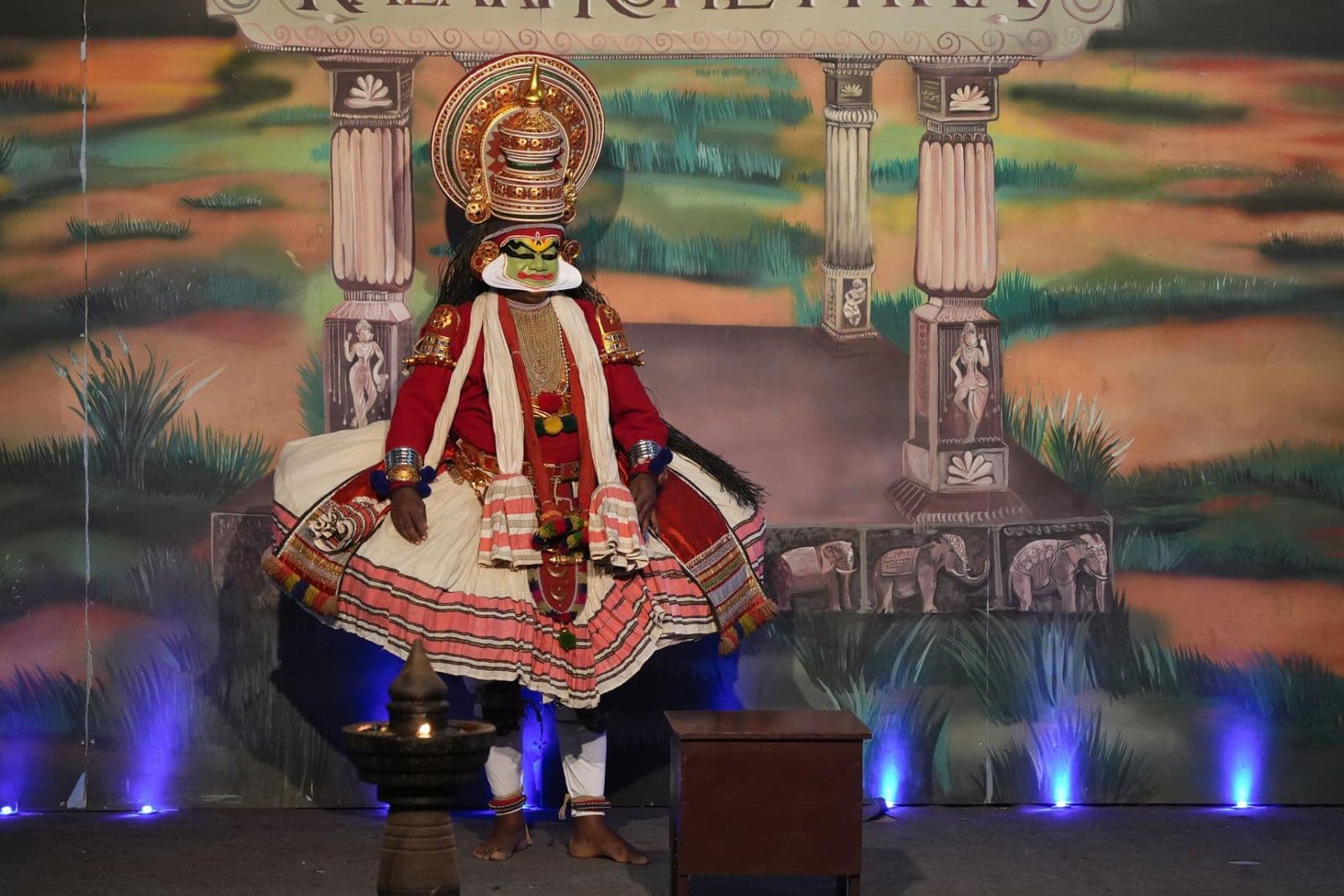 A Kathakali performer in full costume, painted face and headdress, on a lit stage