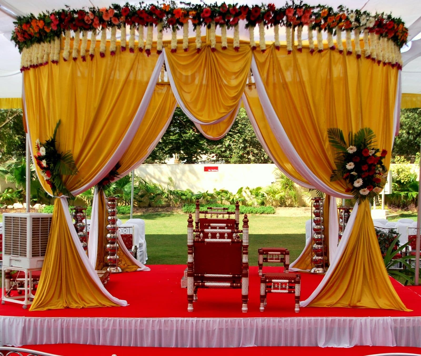 A wedding mandap draped in marigold-yellow fabric with red chairs on a lawn